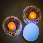 Twee kandelaars van mozaïekglas met gloeiende LED-kaarsen staan op een houten oppervlak naast de Tafellamp DORSETTO oplaadbaar 105 uur, een waterdichte, ovale wit-gouden lamp met afstandsbediening.