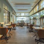 Een helder, modern restaurant met grote ramen, bruine draaistoelen en houten tafels gloeit warm met LED waxinelichtjes 3D (175 uur brandtijd, oplaadbaar, afstandsbediening & timer, set van 12), terwijl gasten buiten genieten van groene planten en uitzicht op bomen.