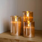 Drie DORSETTO oplaadbare led kaarsen in amberkleurige houders, 105 uur brandtijd en afstandsbediening, staan op een houten plank en geven sfeervol licht. Het magnetisch oplaadstation kleurt rood tijdens het opladen van de 3-delige set.