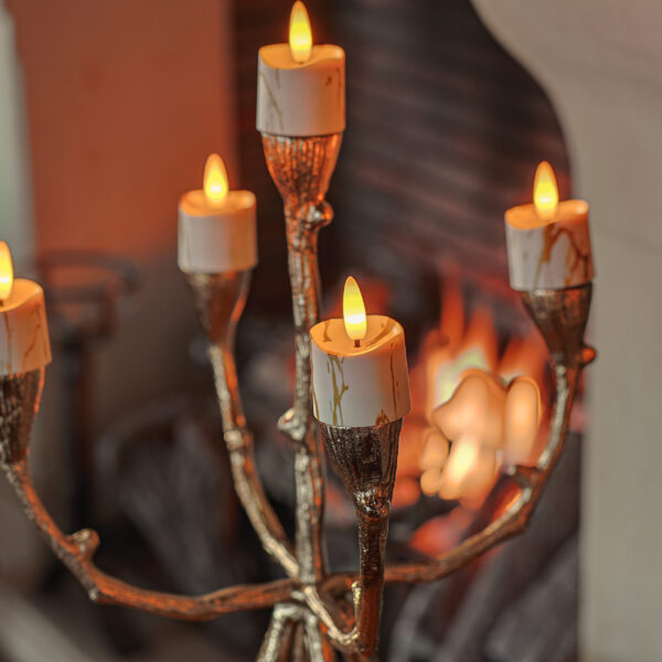 Een gouden kandelaar met vijf witte, brandende kaarsen en Oplaadbare LED waxine/theelichtjes 175 uur waterproof warm wit (6 stuks) met afstandsbediening staat binnen, met een warme haard die zachtjes gloeit op de onscherpe achtergrond.