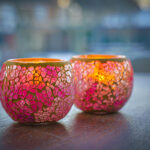 Twee Waxinelichthouder 8,5 CM Dorsetto mozaïek roos kandelaars met roze en oranje mozaïekglas branden op een houten oppervlak en werpen een warme gloed. De zacht vervaagde achtergrond versterkt de gezellige sfeer.