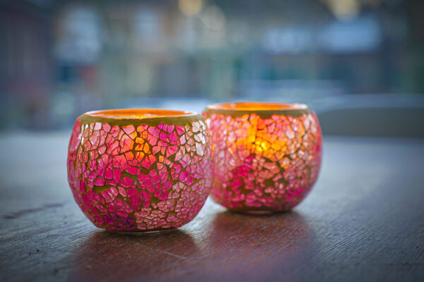 Twee Waxinelichthouder 8,5 CM Dorsetto mozaïek roos kandelaars met roze en oranje mozaïekglas branden op een houten oppervlak en werpen een warme gloed. De zacht vervaagde achtergrond versterkt de gezellige sfeer.