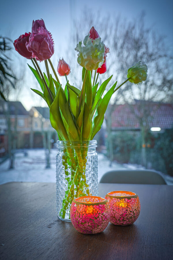 Een heldere glazen pot met roze en witte tulpen staat op een houten tafel naast twee brandende Waxinelichthouder 8,5 CM Dorsetto mozaïek rozenkandelaars. Op de achtergrond staat een besneeuwde tuin met charmante huisjes en bomen.