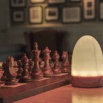 In een warm verlichte kamer staat de tafellamp DORSETTO oplaadbaar 105 uur, waterdicht en ovaal met marmer met grijze houtlook en afstandsbediening, te stralen naast een houten schaakbord met uitgesneden stukken op tafel.