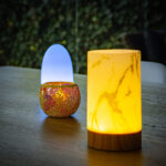 Drie decoratieve lampen rusten op een houten tafel: een lamp met geel marmerdessin aan de voorkant, een lamp met goudstructuur en paars licht zoals de Waxinelichthouder 8,5 CM Dorsetto mozaïekroos in het midden, en een blauwgloeiende lamp aan de achterkant, met groen erachter.