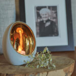 Een LED waxine lichtjes oplaadbaar met afstandsbediening (waterdicht, 175 uur brandtijd) schijnt in een wit-gouden houder op een houten plank, naast witte bloemen. Achtergrond: ingelijst zwart-wit foto van een oudere vrouw.