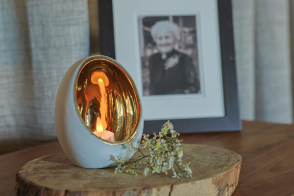 Een LED waxine lichtjes oplaadbaar met afstandsbediening (waterdicht, 175 uur brandtijd) schijnt in een wit-gouden houder op een houten plank, naast witte bloemen. Achtergrond: ingelijst zwart-wit foto van een oudere vrouw.