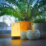 Twee potplanten staan op een vensterbank naast een gloeiende kaars en twee Theelichthouder DORSETTO 9 cm donker groen, met zacht natuurlijk licht door het raam op de achtergrond.