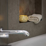 Een brandende kaars in een Theelichthouder DORSETTO 9 cm donker groen en een netjes opgerolde beige handdoek liggen op een verzonken badkamerplank boven het bad, met een chromen kraan zichtbaar op de voorgrond.