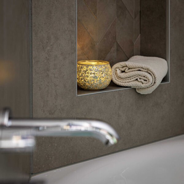 Een brandende kaars in een Theelichthouder DORSETTO 9 cm donker groen en een netjes opgerolde beige handdoek liggen op een verzonken badkamerplank boven het bad, met een chromen kraan zichtbaar op de voorgrond.