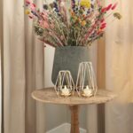 Een rustieke houten tafel met een grote vaas met kleurrijke droogbloemen en een Waxinelichthouderset DORSETTO metaal rosé goud kleurig per 2 stuks, elk met brandende waxinelichtjes, is gedekt tegen beige gordijnen.