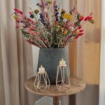 Op een rustieke houten ronde tafel staat een grote vaas met kleurrijke droogbloemen en een Waxinelichthouderset DORSETTO metaal rosé goudig per 2 stuks met kleine brandende kaarsjes, tegen beige gordijnen.