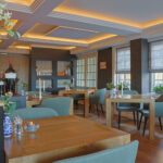 Een stijlvol restaurant met houten tafels, groene stoelen, grote ramen met vouwgordijnen, elegante verlichting en accenten zoals een blauw-witte vaas en oplaadbare LED waxine/theelichtjes 175 uur waterdicht warm wit (6 stuks) met afstandsbediening.