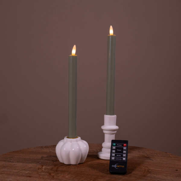 Twee verlichte Dinerkaarsen LED 400 uur brandduur oplaadbaar olijfgroen (set van 2) rusten in witte keramische houders op een houten oppervlak, met een kleine zwarte Doggetto afstandsbediening ervoor. De achtergrond is effen bruin.