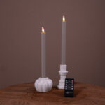 Twee donkergrijze Dinerkaarsen LED-kaarsen (oplaadbaar, brandtijd 400 uur, set van 2) in witte houders staan op een houten oppervlak tegen een bruine achtergrond. Een kleine afstandsbediening van DSETTO is ervoor geplaatst.