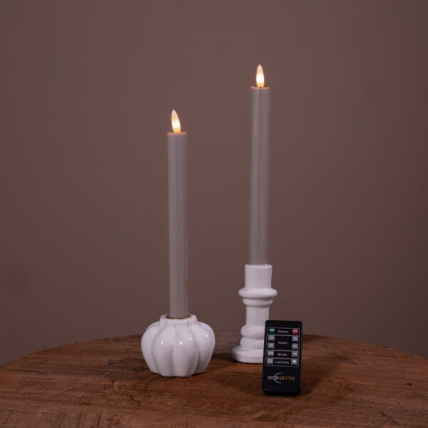 Twee donkergrijze Dinerkaarsen LED-kaarsen (oplaadbaar, brandtijd 400 uur, set van 2) in witte houders staan op een houten oppervlak tegen een bruine achtergrond. Een kleine afstandsbediening van DSETTO is ervoor geplaatst.