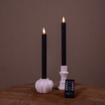 Twee beige oplaadbare LED dinerkaarsen (set van 2, brandduur 400 uur) zitten in witte houders - een pompoenvormig, een cilindervormig - op een houten oppervlak met een afstandsbediening ervoor, tegen een effen achtergrond.