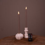 Zwei graue LED-Spitzkerzen in weißen Haltern stehen auf einer Holzfläche, vor denen eine kleine Fernbedienung mit der Aufschrift DESETTO platziert ist. Die Kerzen werden angezündet und schaffen ein warmes Ambiente.