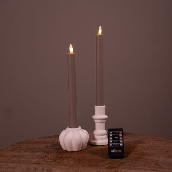 Zwei graue LED-Spitzkerzen in weißen Haltern stehen auf einer Holzfläche, vor denen eine kleine Fernbedienung mit der Aufschrift DESETTO platziert ist. Die Kerzen werden angezündet und schaffen ein warmes Ambiente.