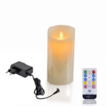 Die wiederaufladbare LED-Wachskerze Ø 7,5 × 15 cm - warmweiß leuchtet flammenlos. Daneben liegen ein schwarzes Netzteil und eine weiße Fernbedienung mit bunten Funktionsstasten.