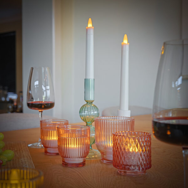 Ein Holztisch mit rosafarbenen Glashaltern mit LED-Teelichtern wiederaufladbar mit Fernbedienung, die warm leuchten. Zwei höhere Kerzenhalter, ein Weinglas mit Rotwein, ein Glaskrug und Weintrauben vervollständigen die einladende Szene.