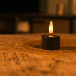 Twaalf Oplaadbare waxinelichtjes zwart met een brandduur tot 400 uur en afstandsbediening staan op een houten oppervlak en werpen een warme gloed. De zacht vervaagde achtergrond creëert een gezellige, ontspannen sfeer.