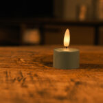 Een set van 12 olivijngroene LED waxinelichtjes met afstandsbediening staat op een houten oppervlak, werpt een warme, zachte gloed voor maximaal 400 uur en creëert een gezellige, intieme sfeer met zijn zachte licht.