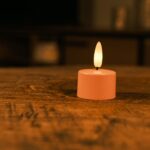 Eine kleine, rosafarbene, batteriebetriebene LED-Kerze mit einer gelben Flamme steht auf einem Holztisch in einem schwach beleuchteten Raum mit unscharfem Hintergrund.