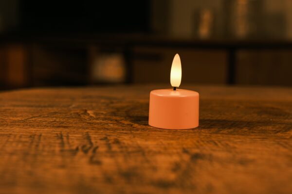 Eine kleine, rosafarbene, batteriebetriebene LED-Kerze mit einer gelben Flamme steht auf einem Holztisch in einem schwach beleuchteten Raum mit unscharfem Hintergrund.