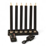 Sechs schwarze elektrische Kerzen mit flammenförmigen Glühbirnen sind in einem rechteckigen schwarzen Halter angeordnet. Davor befinden sich eine schwarze Fernbedienung und ein Steckernetzteil, alles auf weißem Hintergrund.