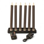 Sechs hohe, graue elektronische Kerzen mit brennenden Spitzen sind in einem schwarzen rechteckigen Halter angeordnet. Vor dem Halter befinden sich eine Fernbedienung und ein schwarzer Stromadapter, alles vor einem weißen Hintergrund.