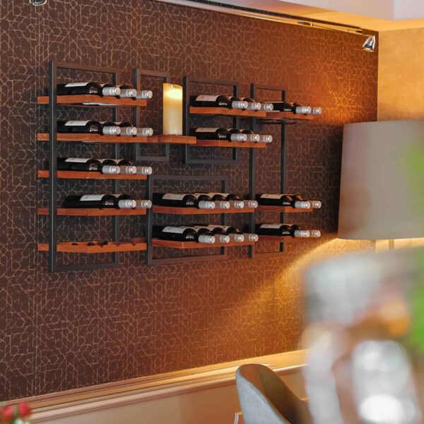 Ein modernes Wandweinregal mit mehreren Fächern fasst mehrere Weinflaschen und die LED-Wachskerze wiederaufladbar DORSETTO D 7,5 × 20 CM, die vor einer gemusterten braunen Wand in einem stilvollen, warm beleuchteten Raum steht.