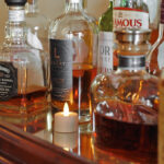 Mehrere Spirituosenflaschen, darunter Cognac und Whiskey, stehen auf einer spiegelnden Fläche neben einem LED-Teelichter wiederaufladbar mit Fernbedienung - wasserdicht - bis zu 400 Stunden Brenndauer.