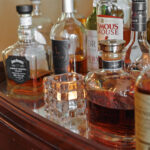 Ein Set aus verschiedenen Spirituosenflaschen und einem verspiegelten Holztablett ist mit einem strukturierten Halter dekoriert, der anstelle einer echten Kerze ein warmweißes, wiederaufladbares LED-Teelicht (bis zu 400 Stunden, 12er-Pack, Fernbedienung) enthält.