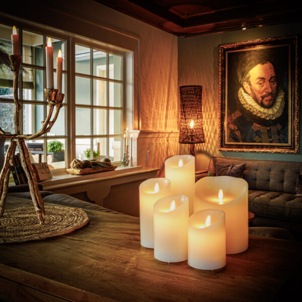 In einem gemütlichen Wohnzimmer leuchten wiederaufladbare LED-Wachskerzen D12,5 × H25 CM auf einem Holztisch, ein Kandelaber mit brennenden Kerzen, ein graues Sofa, ein großes historisches Gemälde und eine warme Raumbeleuchtung.