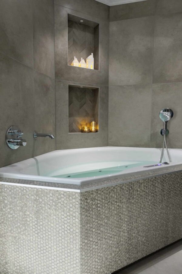 In einer grau gefliesten Ecke steht eine moderne Badewanne mit strukturierten Kacheln. Darüber befinden sich zwei Regale mit wiederaufladbaren LED-Teelichtern (12 Stück, warmweiß, wasserdicht), die mit bis zu 400h Laufzeit und Fernbedienung ein gemütliches Ambiente schaffen.