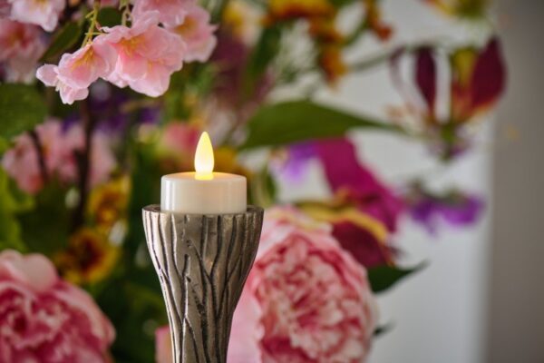 Ein LED-Teelicht wiederaufladbar mit Fernbedienung steht in einem dekorativen Halter vor einem farbenfrohen Strauß pinker und lilafarbener Blumen und sorgt bis zu 400 Stunden lang für eine stimmungsvolle Atmosphäre.