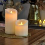 Zwei wiederaufladbare LED-Wachskerzen (D12,5 × H25 cm) mit cremefarbenem Wachs und künstlichen Flammen sitzen auf einer runden Goldmatte auf einem Holztisch. Unscharf leuchtende Kerzen und Vasen schmücken den Hintergrund.