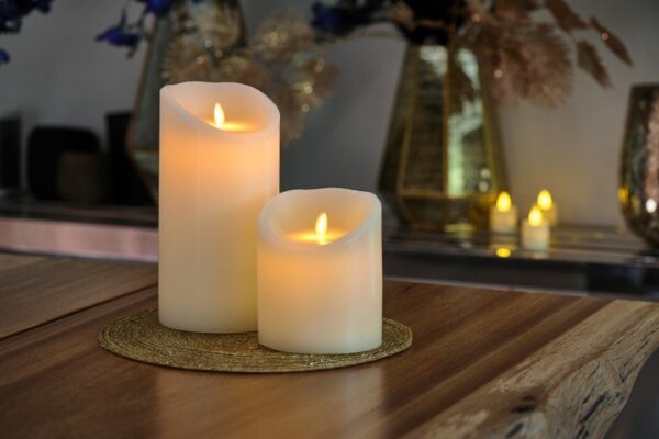 Zwei wiederaufladbare LED-Wachskerzen (D12,5 × H25 cm) mit cremefarbenem Wachs und künstlichen Flammen sitzen auf einer runden Goldmatte auf einem Holztisch. Unscharf leuchtende Kerzen und Vasen schmücken den Hintergrund.