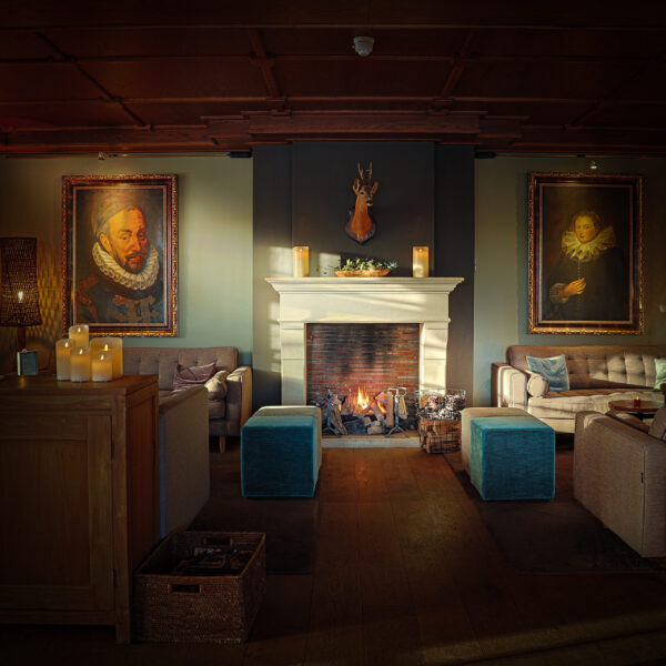 Ein gemütliches, elegantes Wohnzimmer mit einem beleuchteten Kamin, zwei blauen Ottomanen, Sofas und großen gerahmten Porträts am Kaminsims. Die Wärme wird durch die LED-Wachskerze wiederaufladbar DORSETTO D und die Ambientebeleuchtung verstärkt. Über dem Kamin ist ein Hirschkopf angebracht.