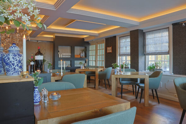 Ein modernes, elegantes Restaurantinterieur mit Holztischen, grünen Stühlen, dekorativen Pflanzen, großen Fenstern mit römischen Jalousien und Deckenleuchten mit warmweißen Wasserfeste LED-Teelichtern (12 Stück) mit Fernbedienung auf den Tischen sorgen für ein gemütliches Licht.